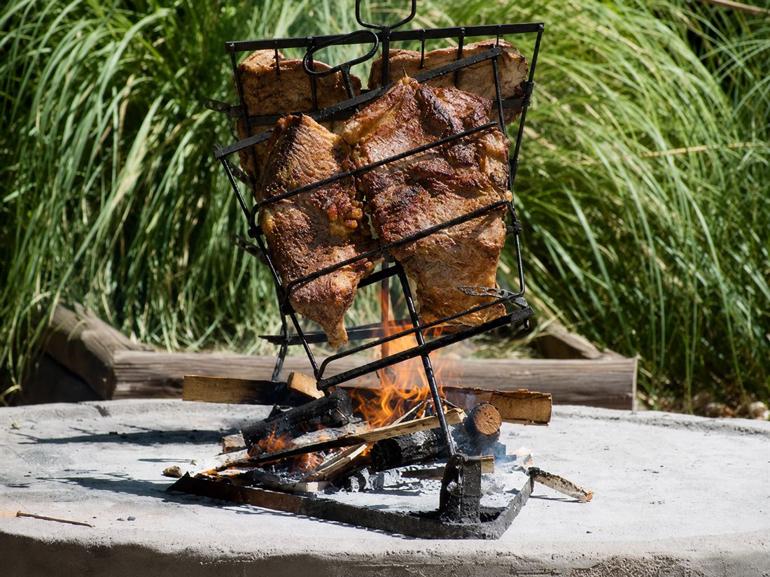 El costillar en cruz ya dorado sobre las llamas, con el pastizal verde detrás