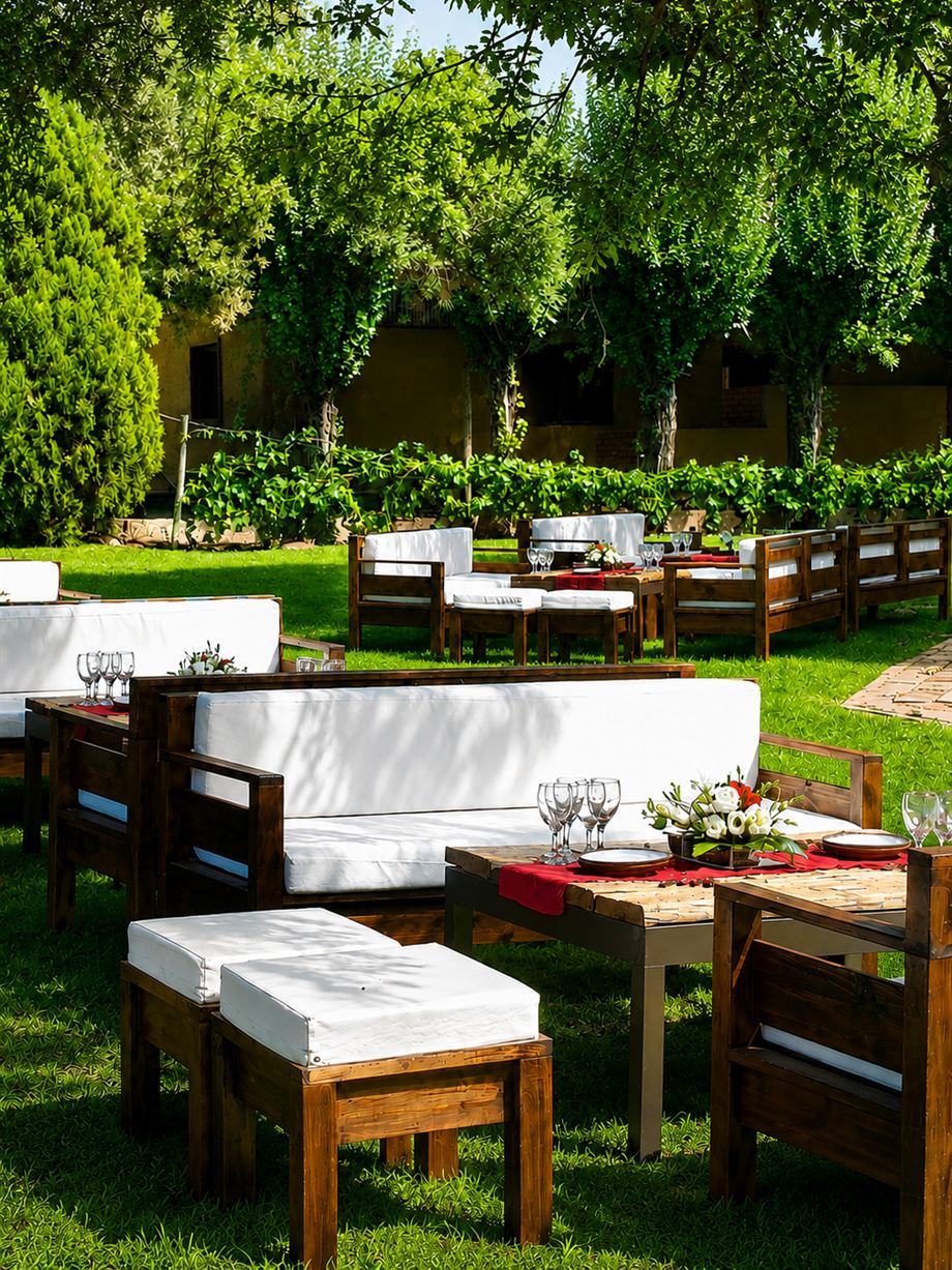 Los juegos de living de madera con almohadones blancos y la mesa tendida con copas y flores, bajo los árboles del parque